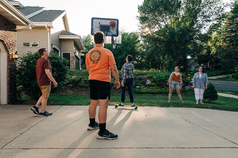 Whole family looks on together as oldest son attempts moving skateboard layup on outdoor basketball hoop in their driveway, "all together now"