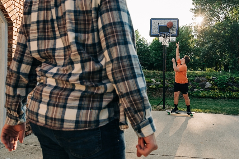 Eldest son in foreground watches as youngest son drains layup while on skateboard in the outdoor basketball hoop in their driveway