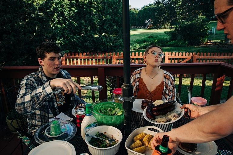 Eldest brother, adult sister and dad select food items for their plates at set patio table with large backyard in the background