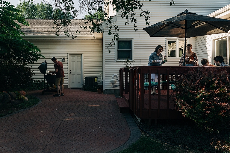 Wide shot: Father arranges "grill charcuterie" while the rest of the family prepares table for the meal at the patio table