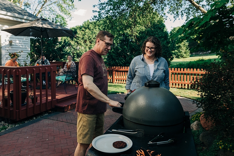 Father and mother work together preparing burger patties on the grill while their three adult children look on from the patio table