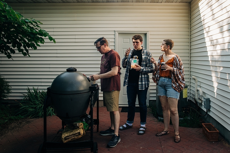 Father works the grill while his adult son and daughter converse and snack on dried fruit and rootbeer