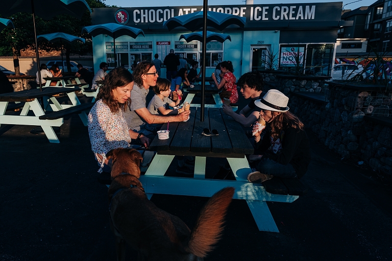 Wide shot: family eats ice cream outside on patio of ice cream shop in the late afternoon during "late spring at home" 