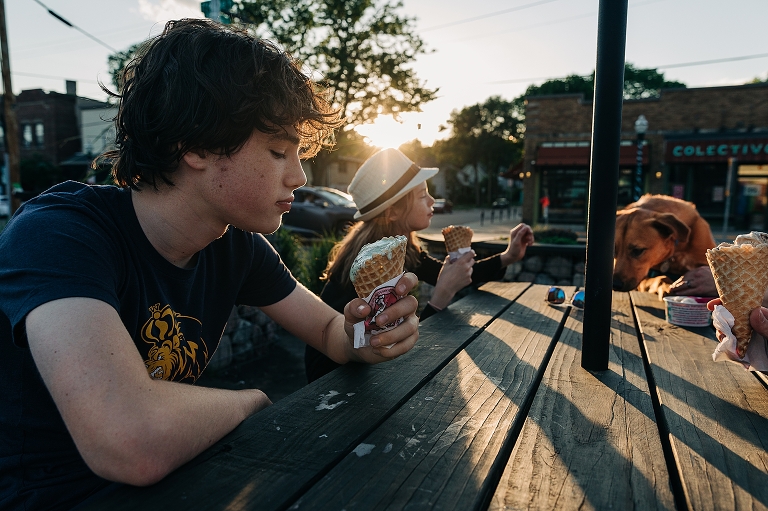 Eldest and middle son enjoy ice cream cones with family dog pawed up on the table in the background in the late afternoon sun