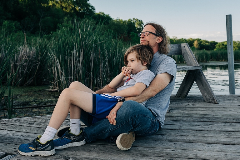Youngest son sits in his father's lap with reeded pond in the background
