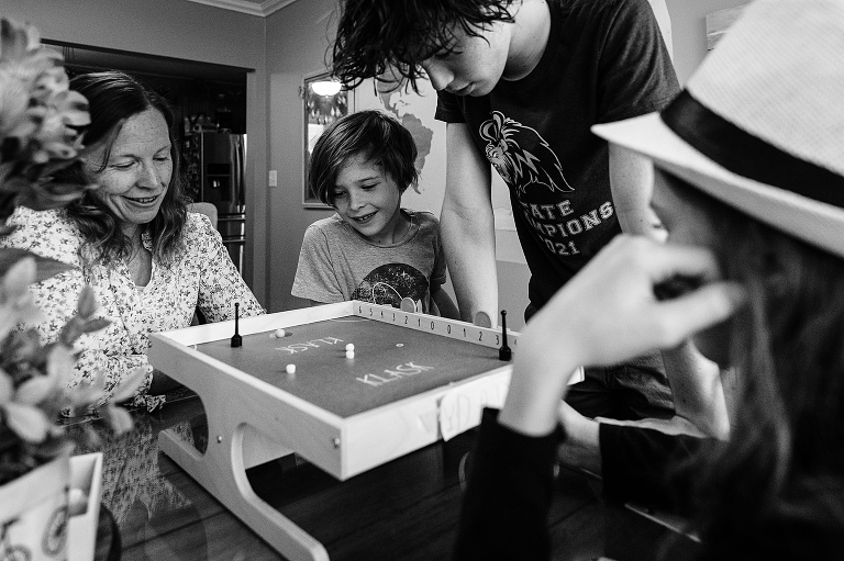 Black and white: Middle son plays klask with mother as youngest and eldest son observe, Middle son in foreground