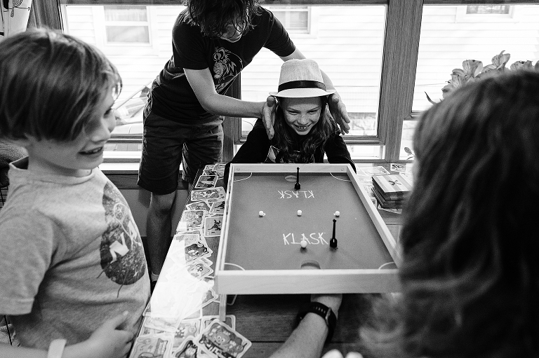 Black and white: Middle son plays klask with mother as youngest and eldest son observe, mother in foreground