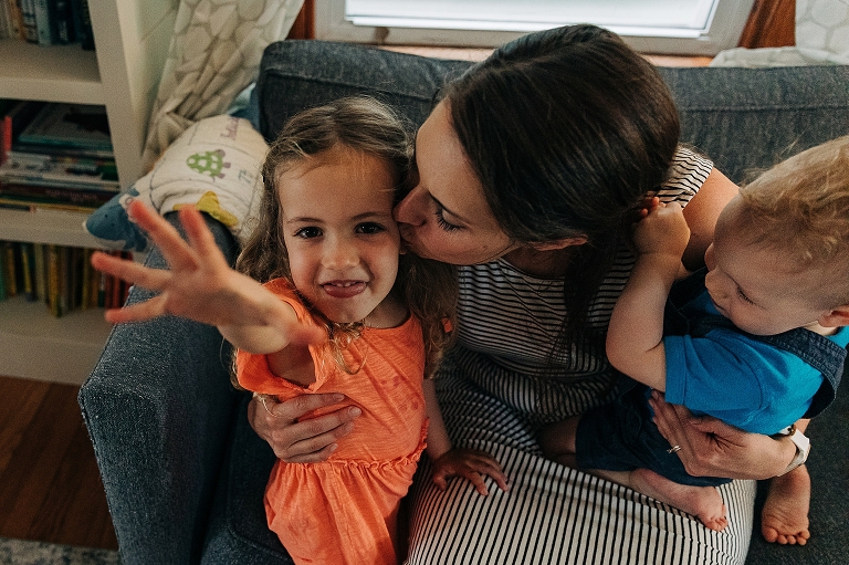 Mother embraces both son and daughter as daughter reaches towards the camera playfully