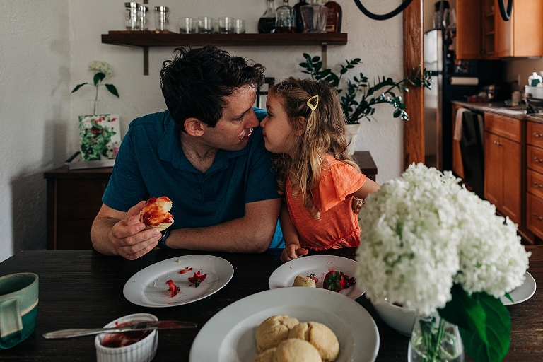 Father in blue shirt and daughter in orange dress make intense/close eye-contact as father eats bagel covered in jam in home