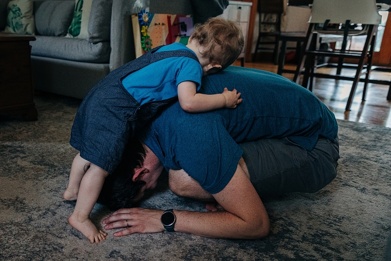 Son in blue overalls hugs father in blue shirt laying in child's pose at home