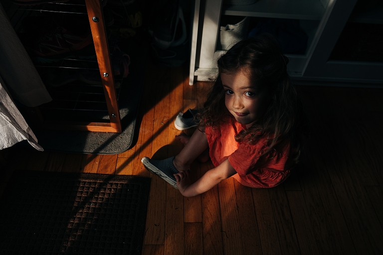 Daughter in orange dress sits in low-light home-hallway smiling