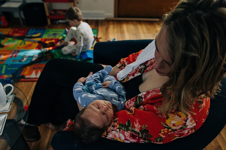 mother holds newborn as young son plays in the background 