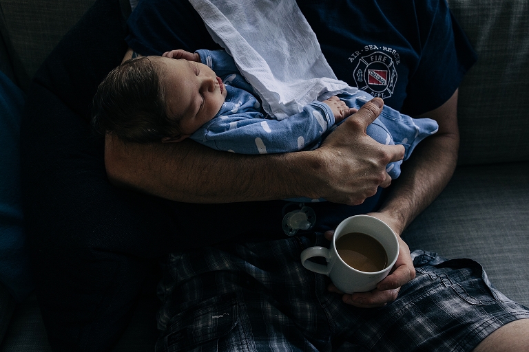 overhead shot of father's lap he holds newborn in one arm and coffee in the other