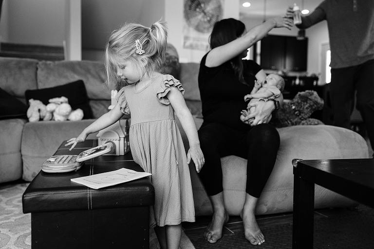 Black and white: Older sister plays with toy keyboard while husband helps mom feed newborn baby in the background