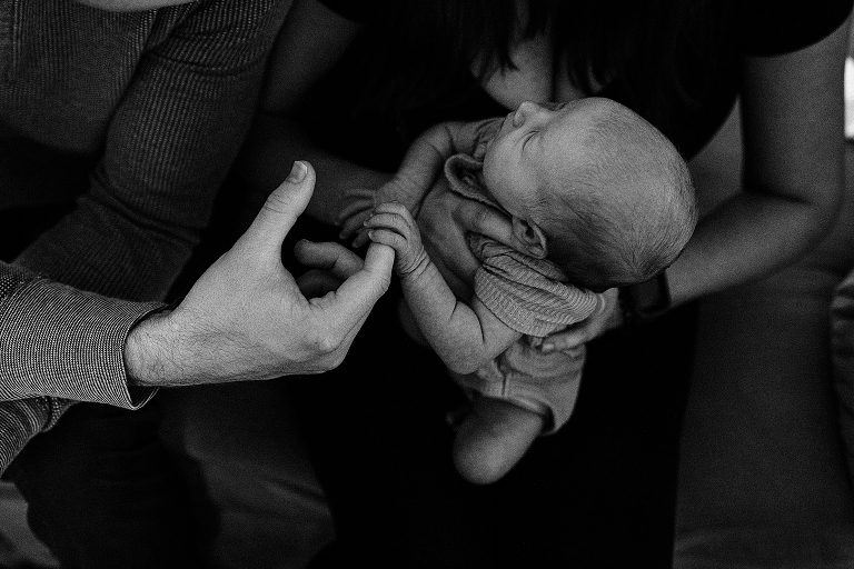 Black and white: Mother holds newborn baby while newborn baby grips fathers pointer finger