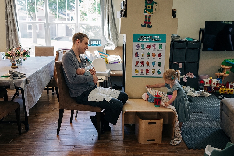 Wide shot: Father holds newborn baby while older sister practices with a doll
