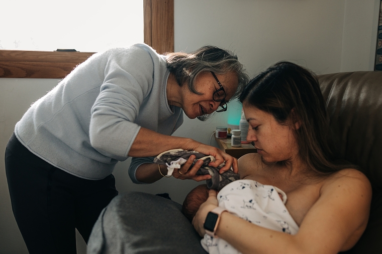 Grandmother helps daughter while she nurses winter newborn in cozy light 