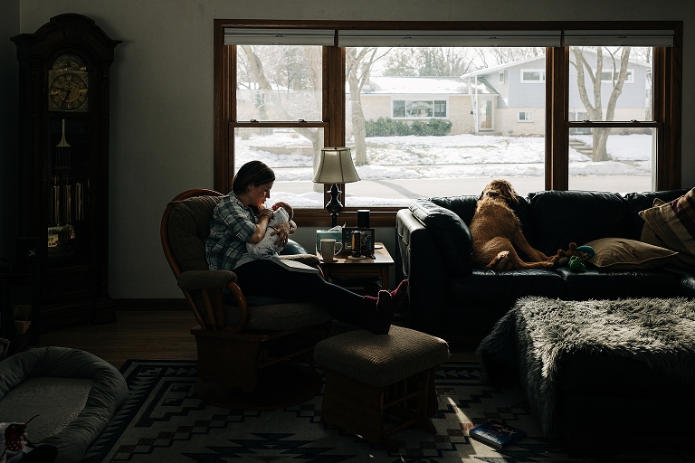 Living room scene of mother holding newborn, dog looking out the window, winter snow on the ground outside and cozy in the home