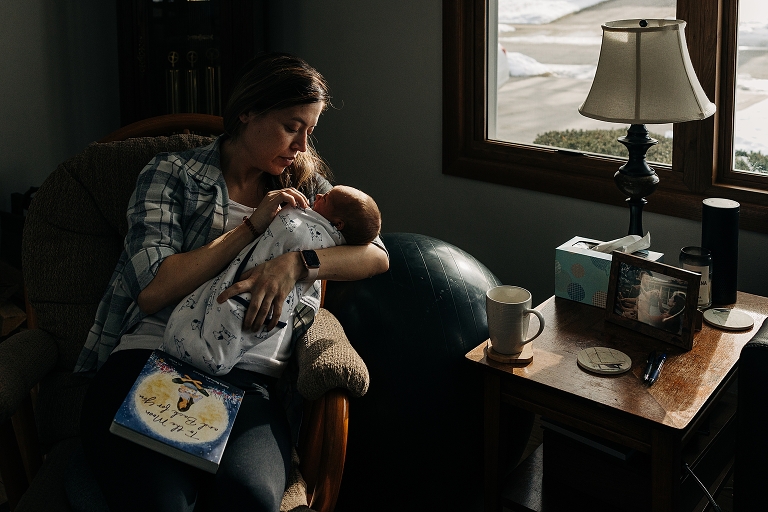 New mother holds newborn in soft winter window light with a book on her lap