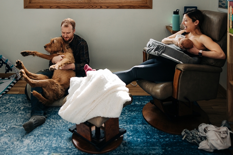 Joyful parents sit on floor and nurse newborn and play with dog on a winter morning in a cozy nursery