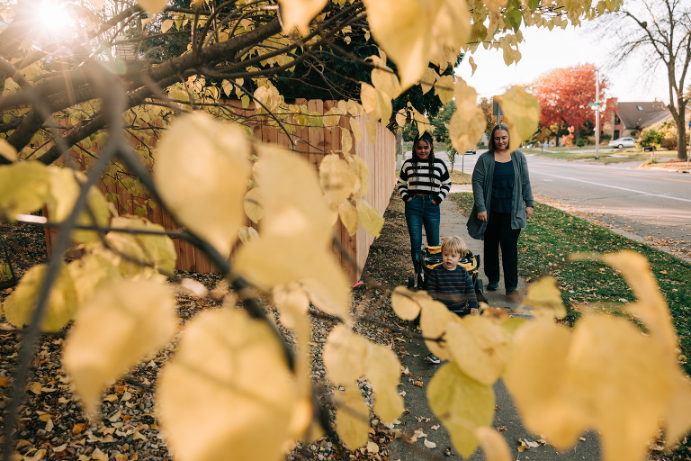 Two women walk behind young boy who is pulling a wagon. The image is framed in yellow fall leaves and golden light coming through the trees.