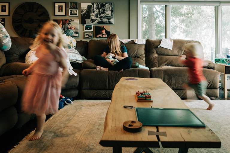 mother nurses newborn while two older children play chase around here. Image of two older children is intentionally blurred to show movement