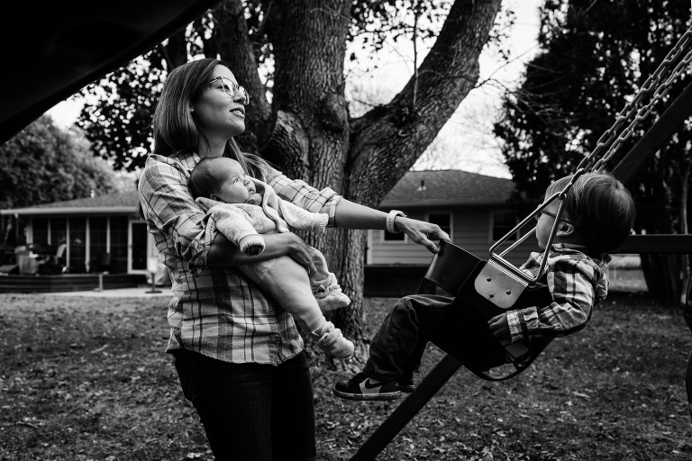mother holds newborn baby whlie helping older child swing in a bucket swing