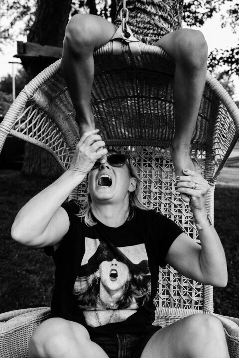 woman sits in an egg chair and playfully pretends to eat her daughter's toes who is sitting above her 