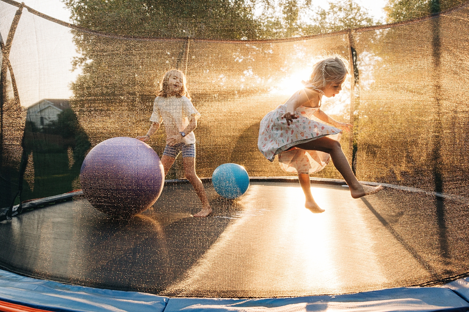 two girls jump on a trampoline on a summer's day. Big beach balls are inside the trampoline. Shadows and light are present