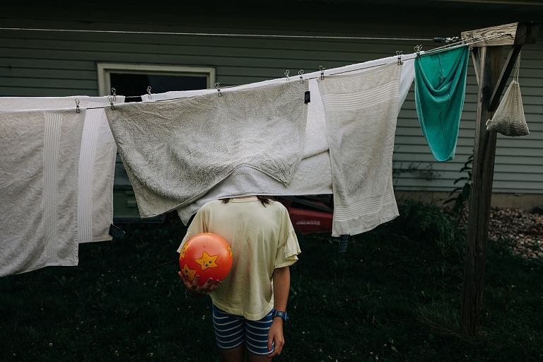 Girl conceals her head with hanging towel while holding ball on end of summer night