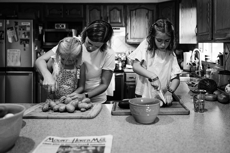 Mother assists youngest daughter in slicing potatoes with knife while eldest slices cucumber by herself