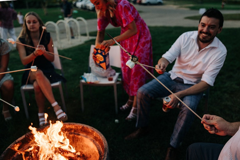 Two men compare roasted marshmallows as wife prepares the next step of the s'mores making process