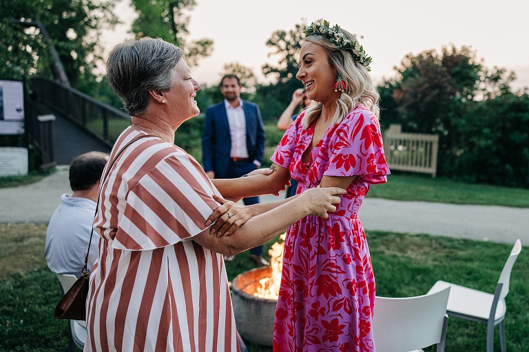 Wife and woman hold hands and exchange frontal conversation with fire pit in the background