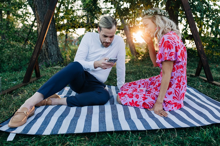 Husband and wife sit on picnic blanket with the sun setting in the background post-ceremony
