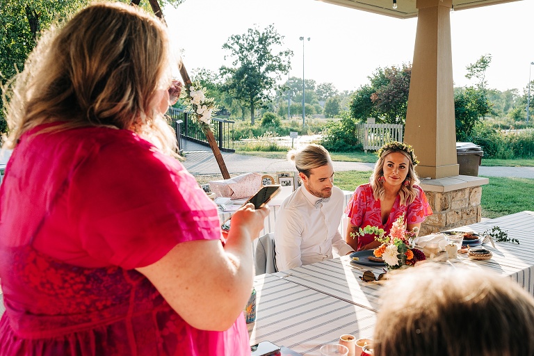 The bridesmaid reads a toast off her phone at Tenney Park Documentary Wedding reception with the newly wed couple watching in the background 