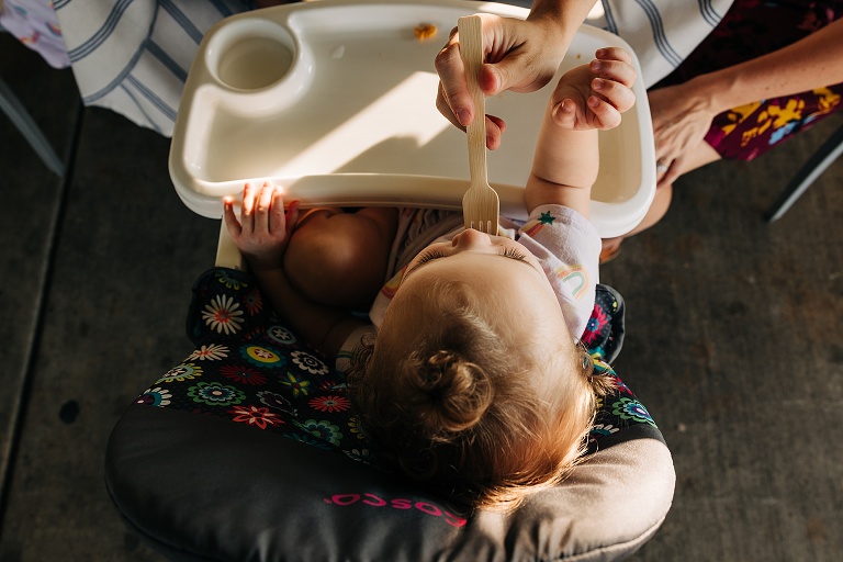 Top down: A toddler in a high chair is fed by a woman with a wooden fork 