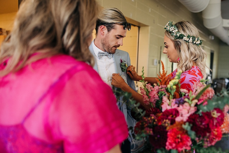 Bride assists groom with corsage with bridesmaid in foreground