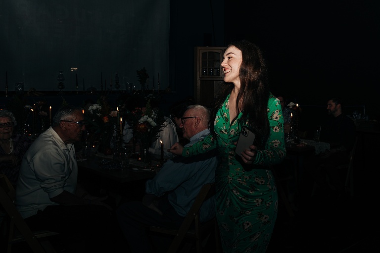 Bridesmaid in green flower pants and long sleeve shirt laughs with male guests conversing in dimly lit background