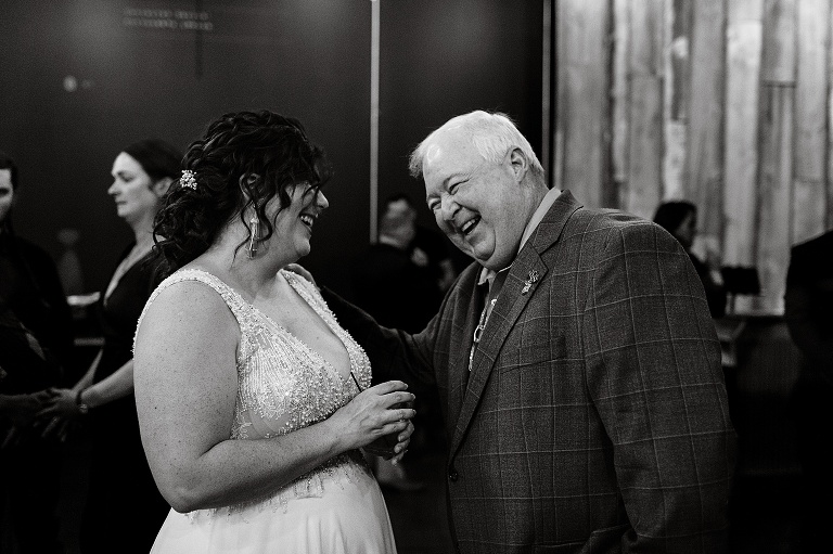 Black and white: father (to wife) and wife laugh with other guests in the background