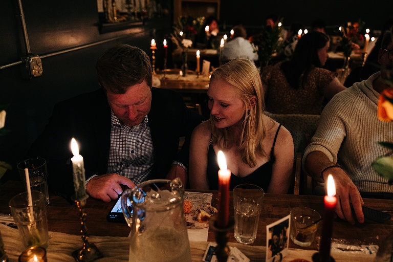 Male and female guest look at male guests phone with interest, lit candles and glasses in the foreground, more lit candles and guests in the background 