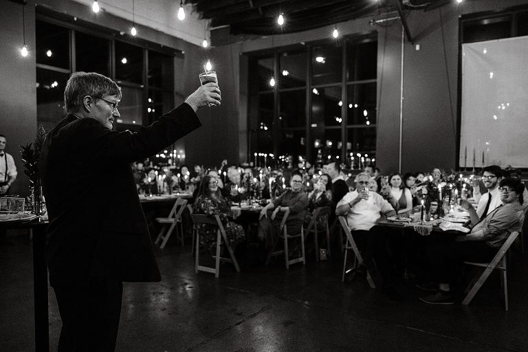Black and white:, wide shot man finishes his toast and raises his glass to the newly wed couple, other guests follow suit