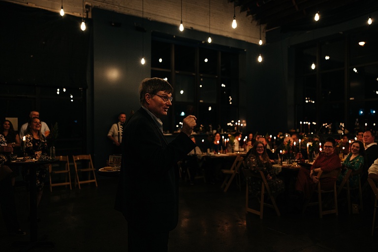 Wide shot: Older man gives toast at restaurant lighted dimly by candles to pleasant reactions from guests