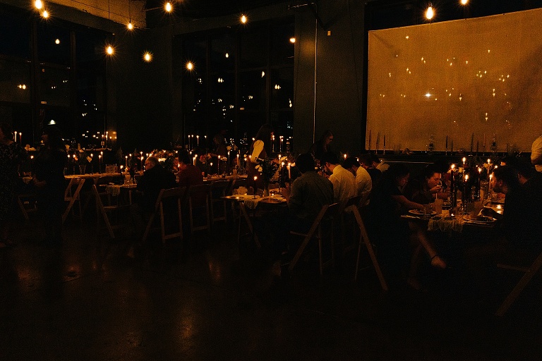 Wide shot: guests sit at the tables of candle lit dim restaurant after Fall Wedding At Home