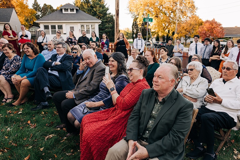 Wide shot: guests react joyfully to the ceremony proceedings with the neighborhood in the background