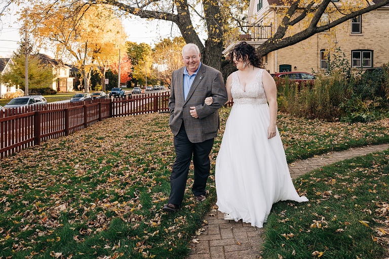 Father of bride walks bride out to the ceremony with a yellow leafed tree in the background