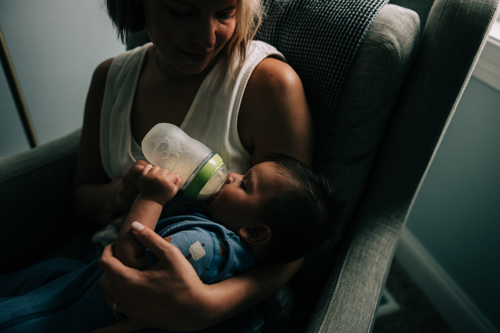 Baby lies in mother's arms taking a bottle in soft window light at the end of the day. Mother looks down at baby, creating a strong composition in beautiful light.